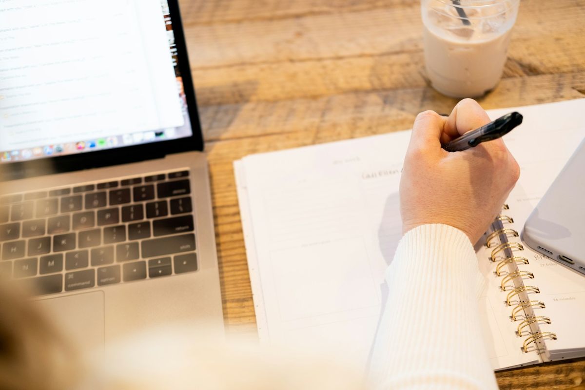 Person noterer arbeidstimer i notatbok ved laptop